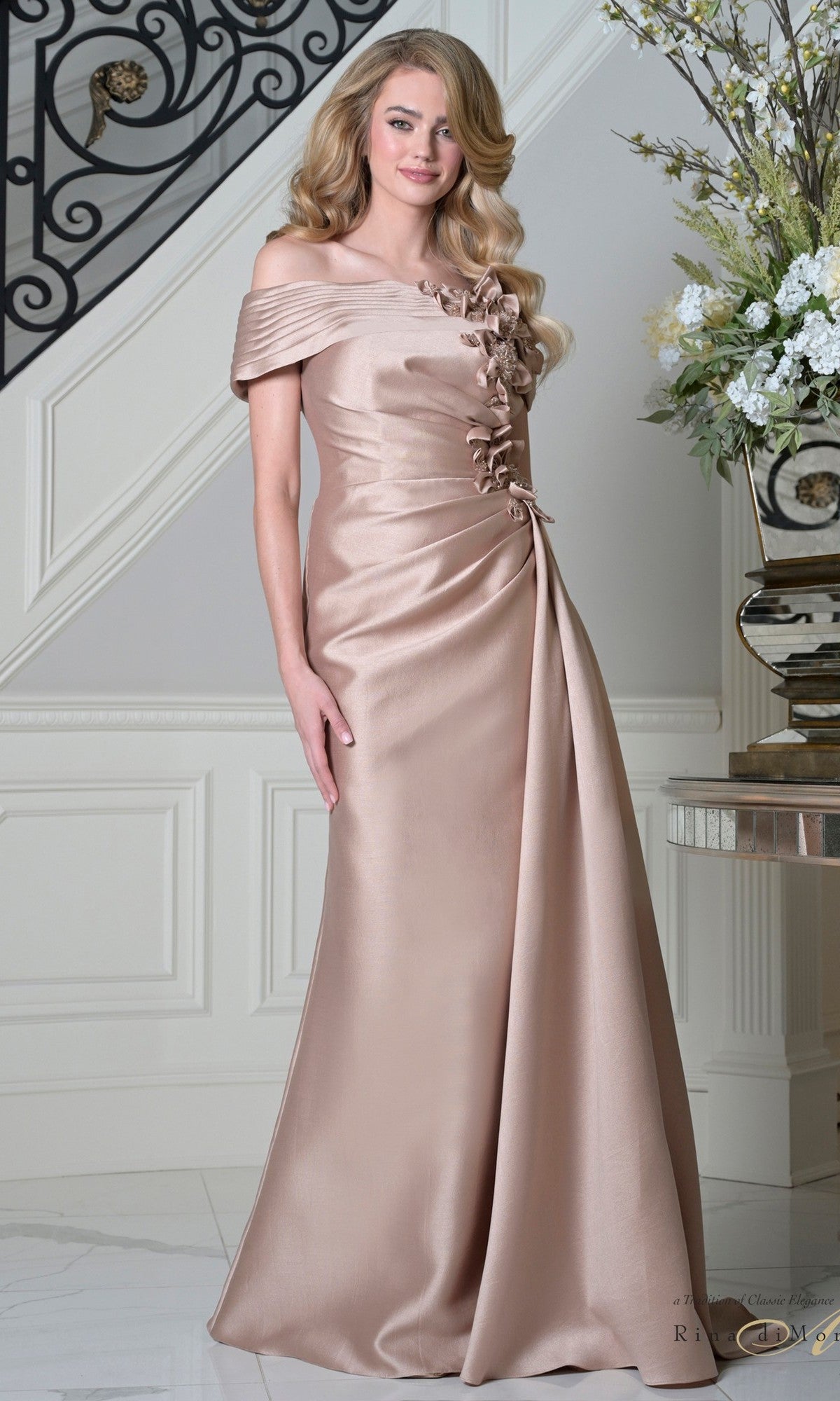 A woman wearing the Rina Di Montella RD3528 long formal dress in rose gold, featuring an off-shoulder neckline and 3D floral appliques, stands indoors near a staircase and a floral arrangement.
